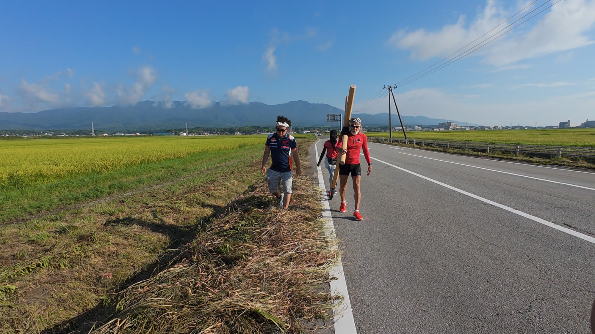 佐渡の稲田と山並みを背に十字架を担ぐアーサー・ホーランドと参加者たち