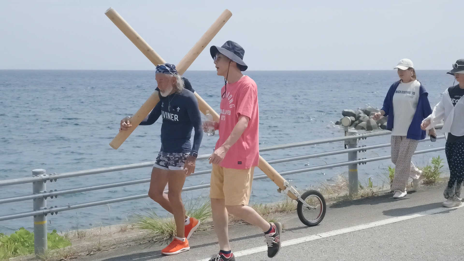 海沿いの道で十字架とともに歩くアーサー先生と同行者たち